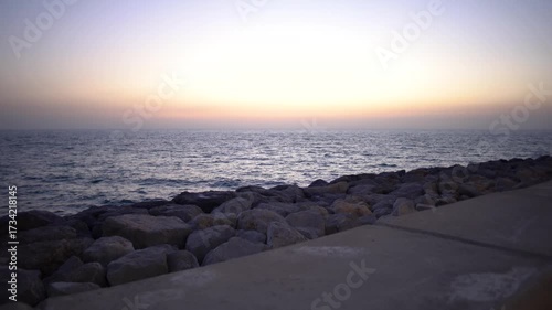 The sky transitions to warm hues as the sun sets beyond the serene ocean. Waves gently lap against a textured rock formation along a peaceful coastal pathway.