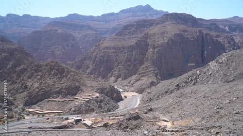A breathtaking view of steep mountain ranges and rocky terrain set in a remote desert area. The sun illuminates the rugged features, showcasing nature's beauty near a winding road.