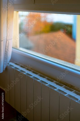 A white central heating radiator under a window, warming a cozy home during the cold autumn season. Concept of home comfort, energy costs, and the start of the heating season.