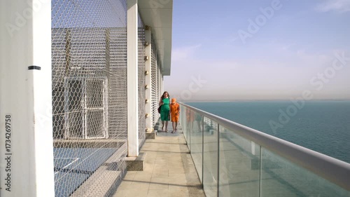A woman strolls along a spacious balcony with a view of the water and city skyline. Bright sunlight illuminates the modern architecture and serene surroundings.