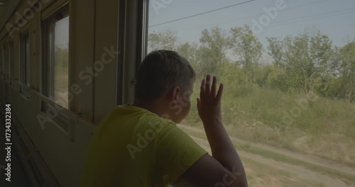 A pensive boy sits by the open window of a passenger train, looking out at the passing landscape of fields and trees during a sunny afternoon ride through the countryside. Rebeok is riding the train.