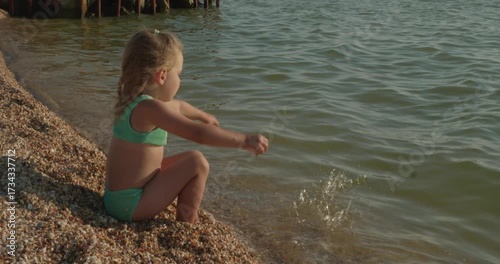 A sweet child squats on a quiet shell beach at sunset, happily collecting and tossing shells into the calm water. The child enjoys a simple, serene moment of his summer vacation at the seaside.
