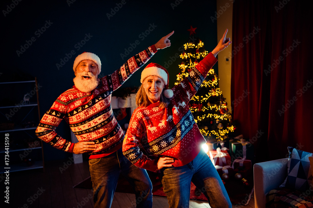 Fototapeta premium Two seniors couple in festive sweaters pose by a Christmas tree at home with gifts and warm lights for a joyful holiday memory