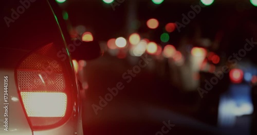 Rear view of car stopped in night traffic, brake light glowing red with stream of blurred headlights and taillights in deep background along crowded road