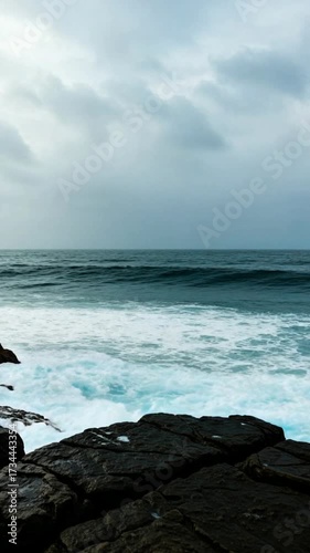 Powerful ocean wave crashes against dark rocks on a cloudy day creating a mesmerizing display of natural force and beauty.
