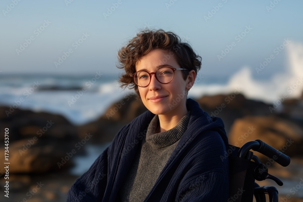 Fototapeta premium Young caucasian female in wheelchair by ocean rocks at sunset