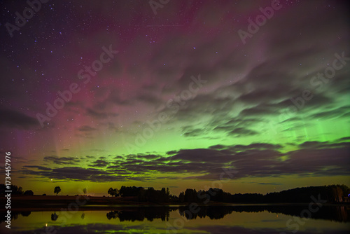 Green and purple aurora lights glow through a starry night sky with scattered clouds, reflecting beautifully on a still lake below.