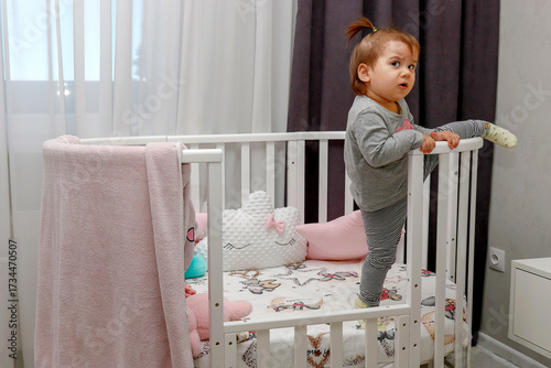 A baby girl tries to climb over the crib railings