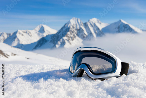 A pair of ski goggles lies on the snow in the mountains against a backdrop of white rocky peaks and a blue sky.