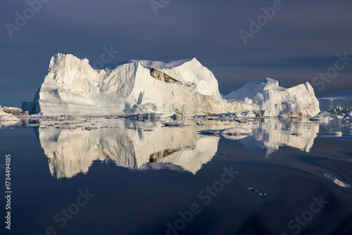 Giant white iceberg floating in calm Arctic waters of Greenland, reflecting perfectly in the sea, under a clear blue sky