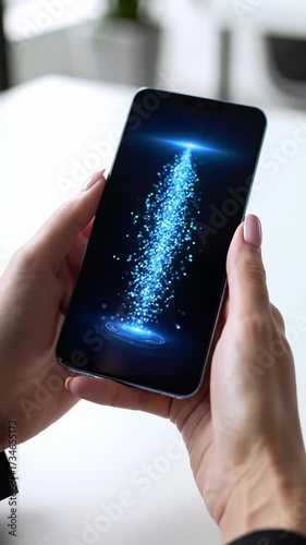 High-angle CU video of clean hands holding a sleek smartphone; blue digital particles stream across the screen. Blurred modern office background. Concept of secure data transfer