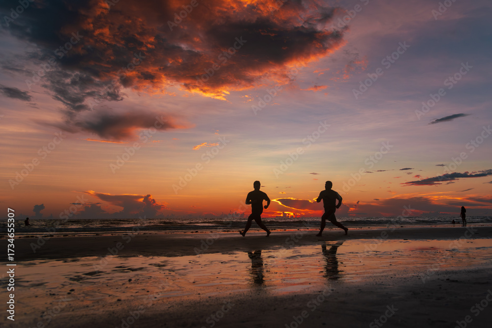 Fototapeta premium Silhouettes of two men running along a beach at sunset