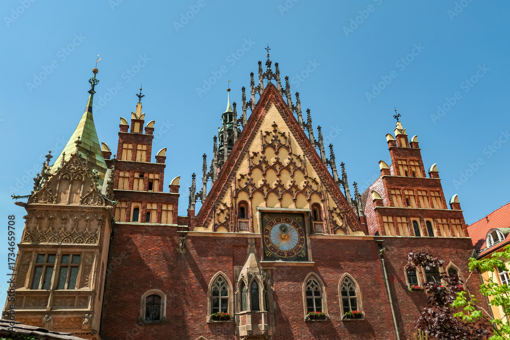 Fototapeta premium The decorative front of Wroclaw Town Hall, with its pointed arches and elaborate clock, stands in bright sunlight, highlighting the intricate details of this historic Polish city landmark.