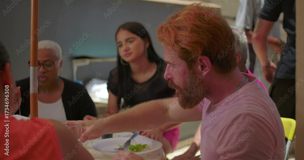 Obraz premium Bearded man drinking water at a backyard BBQ lunch with family and friends, seated at an outdoor table, capturing a relaxed and cheerful gathering in a casual setting