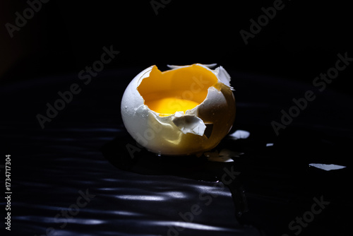 Cracked egg with raw yellow yolk inside the broken shell placed on a dark background. Conceptual food photography symbolizing fragility, new beginnings, nutrition, and culinary preparation.