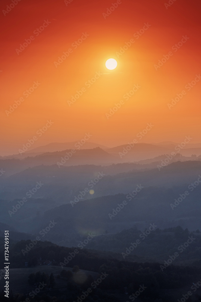 Fototapeta premium Sun setting orange sky above layered rolling hills with atmospheric haze. Capturing serenity and natural beauty at dusk