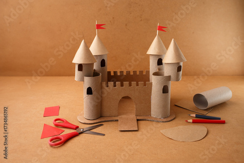 Handmade cardboard castle with red paper flags and crafting tools on table. DIY art project made of paper rolls and recycled materials. Studio composition for design and print. Top view,