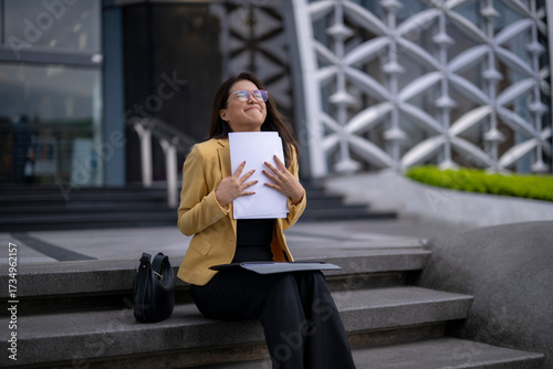 Happy businesswoman holding documents and sitting on stairs outside office building