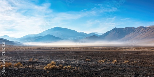 A vast, barren landscape with mountains in the distance, a thin layer of fog or mist in the foreground, and a clear blue sky above.