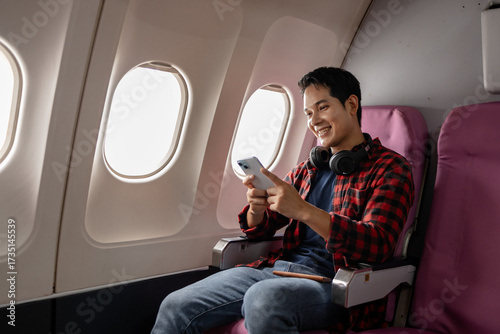 Asian man sitting comfortably on airplane seat near window, holding smartphone and passport, wearing headphones around neck, enjoying in-flight entertainment. Modern travel lifestyle and technology.
