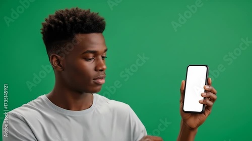 Young Black man holding a smartphone with a blank white screen against a vibrant green background, looking intently at the device.