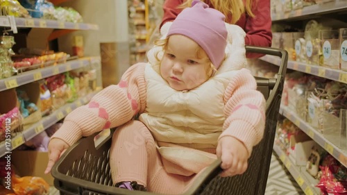 A young child in a pink coat and purple hat sits in a shopping cart as her caregiver pushes her through a grocery store filled with various products. The cheerful atmosphere of the outing is clear.
