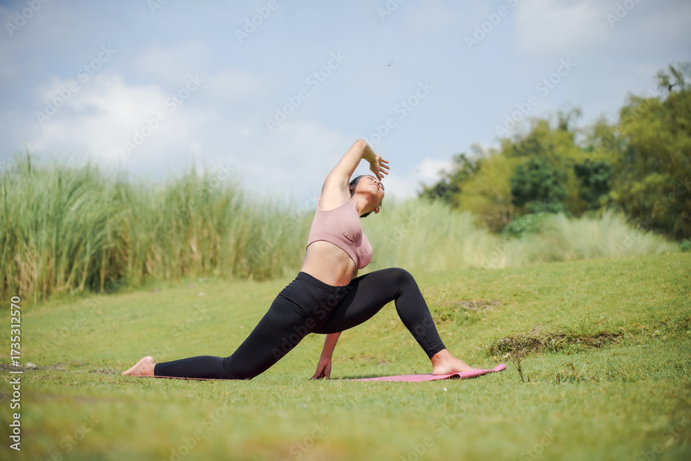 Fototapeta premium Mental and physical health. A slender, tanned Asian woman in sportswear performs yoga outdoors in a beautiful riverside setting in the morning. The woman is performing the Extended Side Angle Pose.
