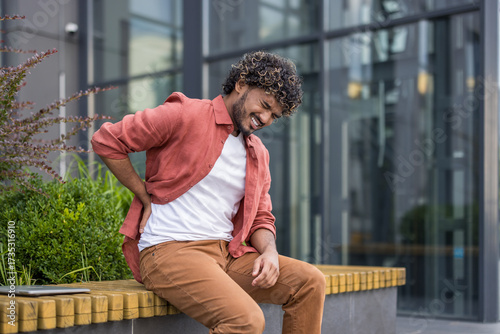 A young Indian man sits on a bench near a building, holding his back with his hand, feeling severe pain