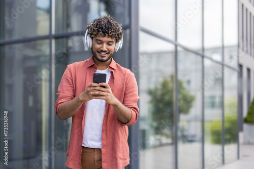 A young Indian man walks through the city wearing headphones and using a mobile phone