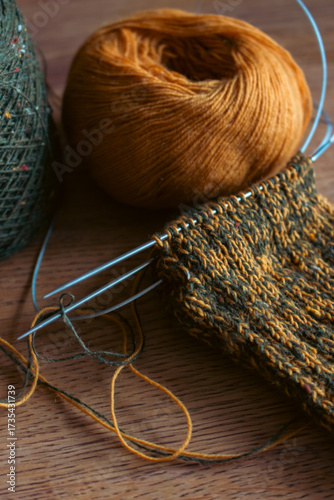 Cozy knitted socks with yarn ball and knitting needles. Knitting process of wool socks. Cozy autumn vibes. Yellow and green tweed yarn on wooden table. Knitting hobby. Knitwear concept. 