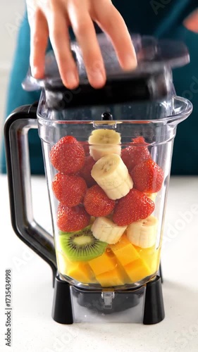 Colorful fruits in blender with hand closing lid in bright kitchen