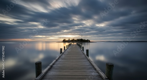 Wooden pier extending towards a small island with reflective water under a dramatic cloudy sky