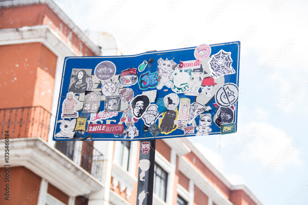 Fototapeta premium Colorful stickers plastered on a public sign, set against a historic Mexican colonial building, highlighting the vivid contrast between street art and traditional architecture.