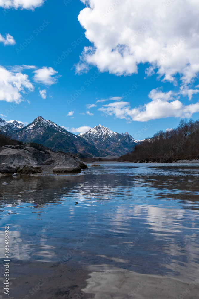 Fototapeta premium Clear Lech River reflecting snowy mountains and clouds near Füssen, Bavaria, Germany – tranquil alpine landscape with blue sky and crystal water