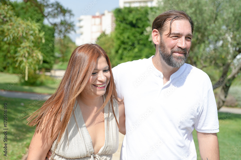 Fototapeta premium A man and a woman are walking together in a park. The man is wearing a white shirt and the woman is wearing a white dress. They are smiling and seem to be enjoying each other's company