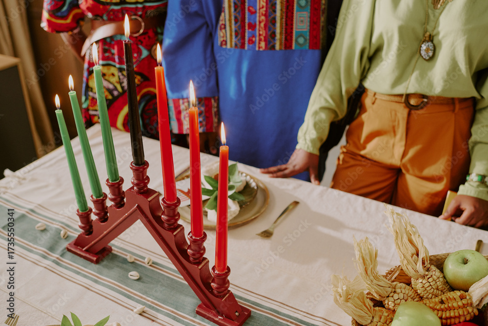 Fototapeta premium Three people standing around table with kinara holding seven candles, celebrating Kwanzaa, visible hands and torsos suggesting young adult and middle aged Black women and man