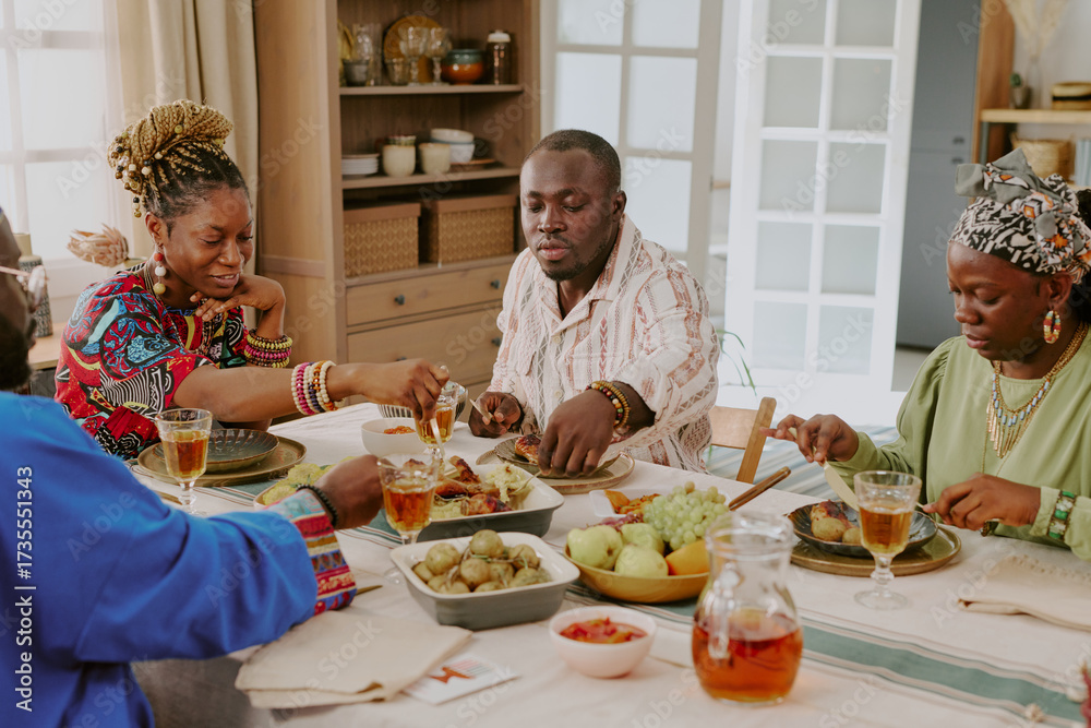 Fototapeta premium Group of Black adults and young adult woman sitting at dining table sharing traditional meal during Kwanzaa celebration, reaching for food and engaging in festive gathering