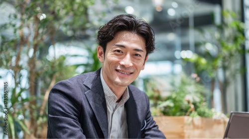A young man dressed in formal attire sits at a desk in a contemporary office. He smiles warmly while surrounded by lush greenery and natural light coming through large windows.