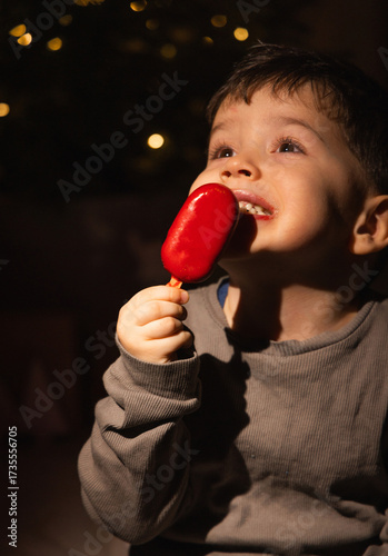Little boy near a decorated Christmas tree holds a fake ice cream shaped like a Santa boot or gingerbread. Child in pajamas tastes sweets and licks fingers in cozy dim light festive New Year atmospher