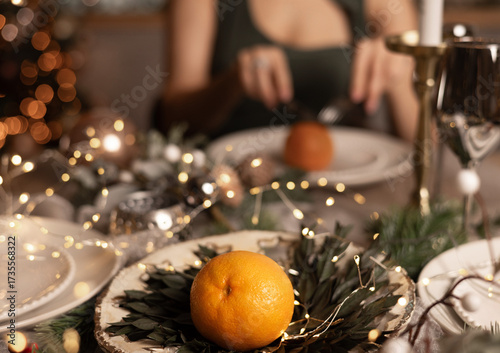 woman sits at a cozy Christmas dinner table decorated with candles and lights. A tall fir tree with globes and toys is behind.girl, female holds a glass of champagne and pretends to eat a mandarin.
