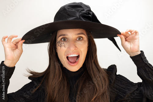 Woman wearing a large black witch hat posing for Halloween. Female with spider web makeup near eyes points to empty space or holds a phone with green screen, showing surprise and smiling.
