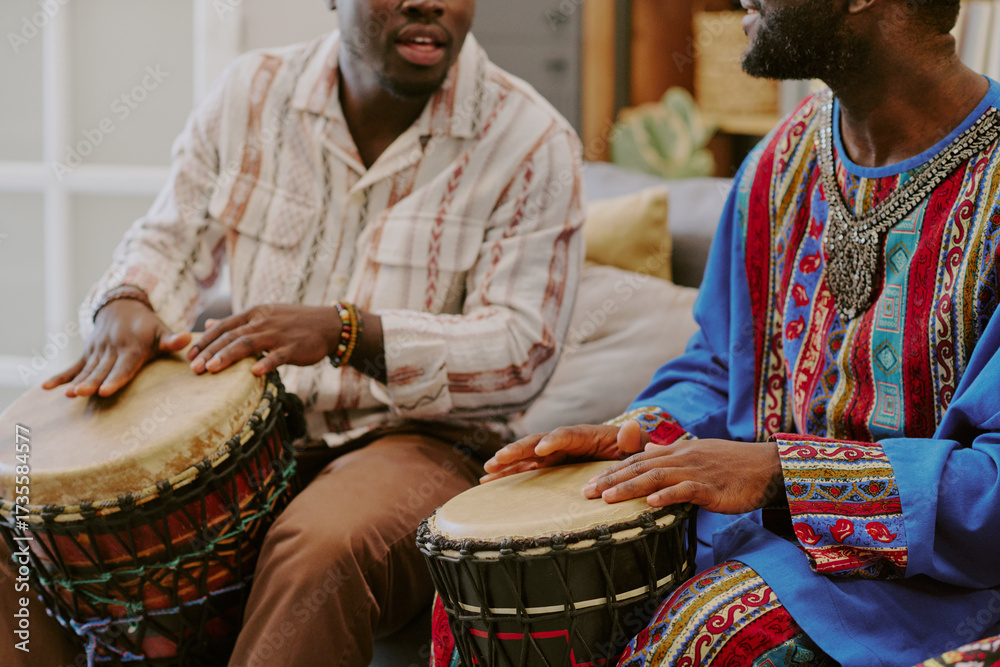 Fototapeta premium Two Black men playing traditional drums during Kwanzaa celebration, sitting side by side, focusing on rhythmic hand movements, wearing patterned clothing, engaging in cultural activity