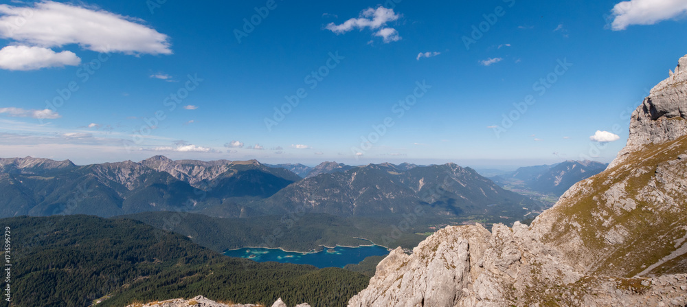 Obraz premium High alpine cliffside view overlooking Eibsee with deep green forests and distant mountain ranges, dramatic panorama near the Zugspitze under clear blue sky