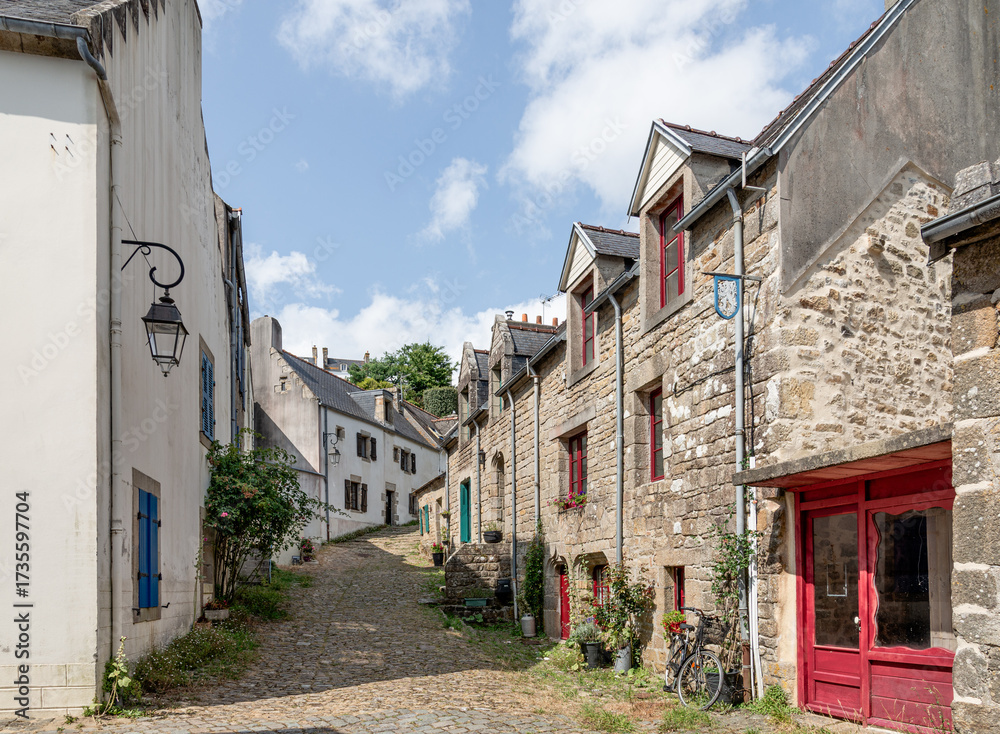 Fototapeta premium A sunlit cobblestone street winds between ancient stone houses, lush greenery, and blooming plants in Pont-Croix in Brittany, France.