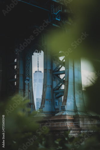 View of the iconic One World Trade Center tower piercing through the steel latticework of the Manhattan Bridge, framed by vibrant green foliage, New York, New York, United States.