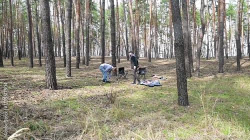 Two men prepare a campfire in a serene pine forest, enjoying an outdoor adventure amidst tall trees and natural beauty. Perfect for themes of camping, friendship, and nature exploration.