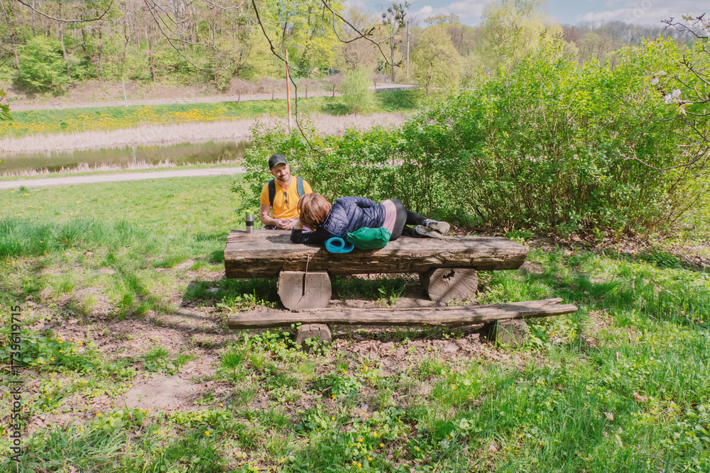 Obraz premium Couple relaxing on a rustic wooden bench in a sunny park with a pond and lush greenery. Perfect for depicting leisure, outdoor adventures, and serene nature breaks.