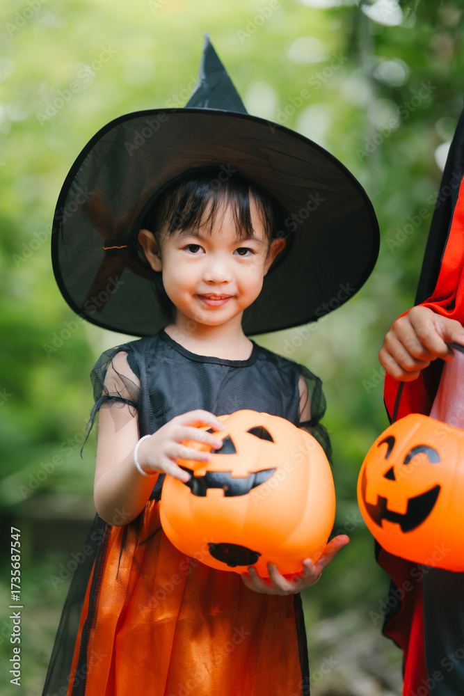 Fototapeta premium A cheerful young girl dressed in a Halloween witch costume smiles happily outdoors. Wearing a pointed black hat and orange dress, she holds a pumpkin jack-o’-lantern bucket, surrounded by greenery