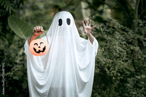 A young boy dressed as a ghost for Halloween wears a white sheet with black eye holes. Holding a pumpkin jack-o’-lantern bucket, he stands outdoors surrounded by dark green foliage