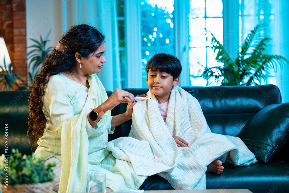 Obraz premium Indian Mother taking son’s temperature while sitting on sofa, showing love and parental care